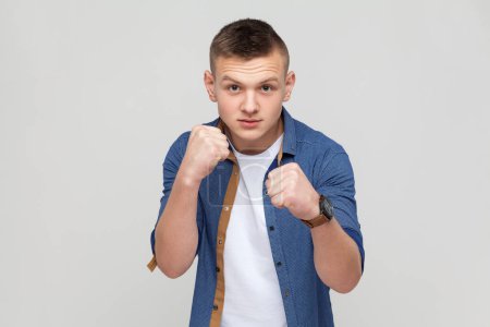 Téléchargez les photos : Portrait of teenager boy in blue shirt standing with boxing fists and ready to attack or defence, looking with angry face, looking at camera with aggression. Indoor studio shot isolated on gray background - en image libre de droit