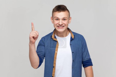 Photo for Portrait of excited amazed teenager boy wearing blue shirt pointing finger up with genius idea, surprised by suddenly invented smart solution. Indoor studio shot isolated on gray background. - Royalty Free Image