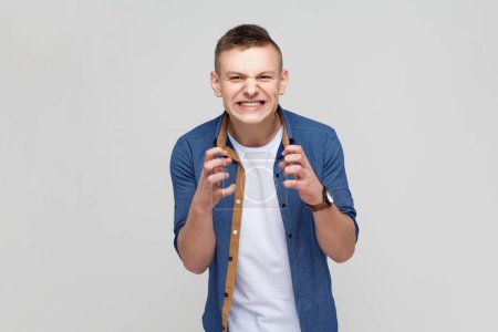 Téléchargez les photos : Portrait of angry nervous teenager boy wearing blue shirt loudly yelling opening mouth holding hands on face, screaming, raised arms. Indoor studio shot isolated on gray background. - en image libre de droit