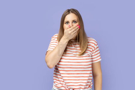 Foto de Portrait of confused shocked blond woman wearing striped T-shirt covering her mouth with palm, don't tell a secret to anyone. Indoor studio shot isolated on purple background. - Imagen libre de derechos