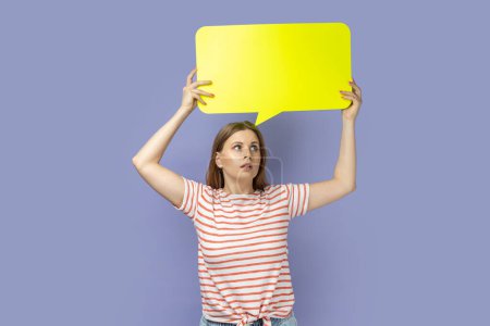 Téléchargez les photos : Portrait of pensive blond woman wearing striped T-shirt looking away and holding yellow bubble thought over her head, copy space for text. Indoor studio shot isolated on purple background. - en image libre de droit