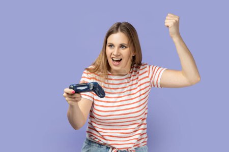 Photo for Portrait of excited amazed blond woman wearing striped T-shirt holding in hands gamepad joystick, clenched fist, showing yes gesture, winning. Indoor studio shot isolated on purple background. - Royalty Free Image