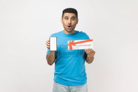 Foto de Portrait of shocked astonished man wearing T- shirt standing with gift voucher and showing smart phone with blank screen, copy space for advertisement. Indoor studio shot isolated on gray background. - Imagen libre de derechos