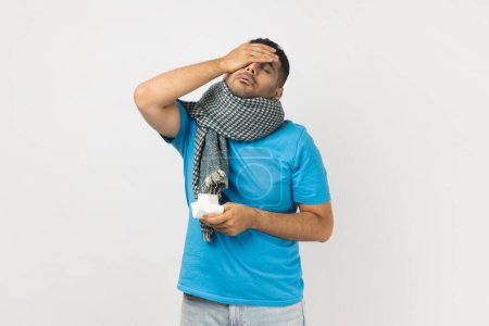 Téléchargez les photos : Portrait of sick ill unheatlhy unshaven man wearing blue T- shirt and wrapped in warm scarf standing touching forehead, has headache, has flu symptoms. Indoor studio shot isolated on gray background. - en image libre de droit