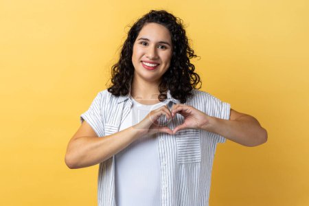 Téléchargez les photos : Portrait of woman with dark wavy hair holding hands in shape of heart showing romantic gesture, love confession, valentines day celebration. Indoor studio shot isolated on yellow background. - en image libre de droit