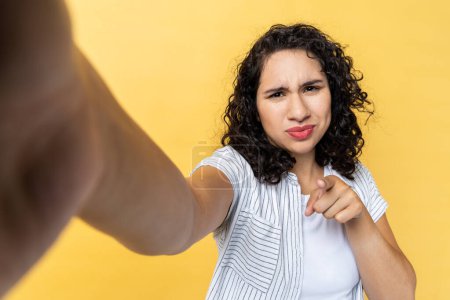 Téléchargez les photos : Beautiful young adult woman blogger with dark wavy hair making point of view photo or broadcasting livestream, pointing finger to camera, POV. Indoor studio shot isolated on yellow background. - en image libre de droit