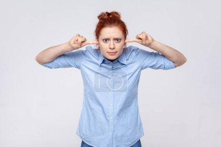Photo for Don't want to listen. Portrait of annoyed redhead woman wearing blue shirt irritated by loud noise covering ears and grimacing in pain. Indoor studio shot isolated on gray background. - Royalty Free Image