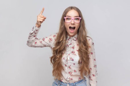 Photo for Portrait of inspired woman in glasses with long blond hair pointing finger up and looking amazed about sudden genius idea, got solution. Indoor studio shot isolated on gray background. - Royalty Free Image