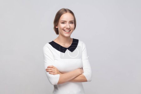 Photo for Portrait of smiling friendly woman wearing white dress standing with crossed arms, looking at camera with toothy smile, expressing positive emotions. Indoor studio shot isolated on gray background. - Royalty Free Image