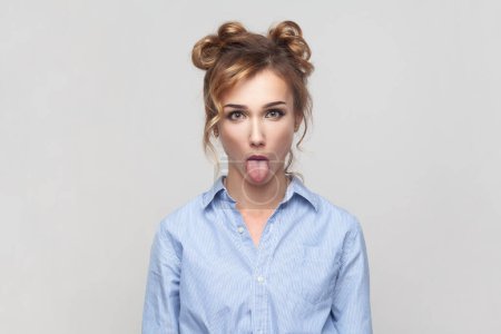 Foto de Portrait of naughty comic blonde woman sticking out tongue, misbehaves, demonstrating her disobedience and protest, has funny look, wearing blue shirt. Indoor studio shot isolated on gray background. - Imagen libre de derechos