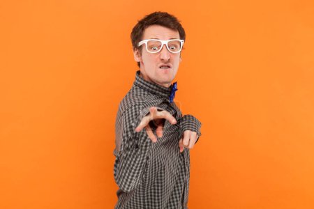 Foto de Retrato de loco divertido hombre discapacitado nerd señalando y mirando a la cámara con los dientes apretados, usando camisa con pajarita azul y gafas blancas. Estudio interior plano aislado sobre fondo naranja. - Imagen libre de derechos