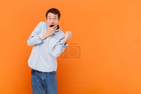 Téléchargez les photos : Man looks with jaw dropped out at camera, being horrified to see something, indicates with thumb aside, advertisement area, wearing light blue shirt. Indoor studio shot isolated on orange background. - en image libre de droit