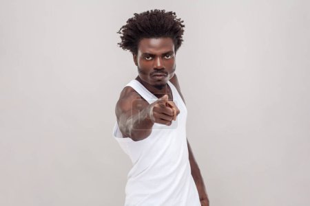 Téléchargez les photos : Portrait of displeased man with Afro hairstyle looks angrily or with judgemental, pointing index finger at camera. wearing white T-shirt. Indoor studio shot isolated on gray background. - en image libre de droit
