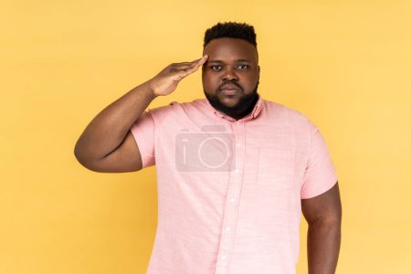 Foto de Yes sir. Portrait of serious man wearing pink shirt holding hand near head as soldier and saluting, looking attentive and confident. Indoor studio shot isolated on yellow background. - Imagen libre de derechos