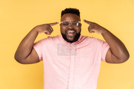Photo for Portrait of delighted satisfied man wearing pink shirt standing, pointing at his new optical spectacles, being happy to see better, smiling happily. Indoor studio shot isolated on yellow background. - Royalty Free Image