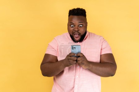 Téléchargez les photos : Portrait of shocked astonished man reading shocking post on social network using cell phone, looking at display with big eyes. Indoor studio shot isolated on yellow background. - en image libre de droit