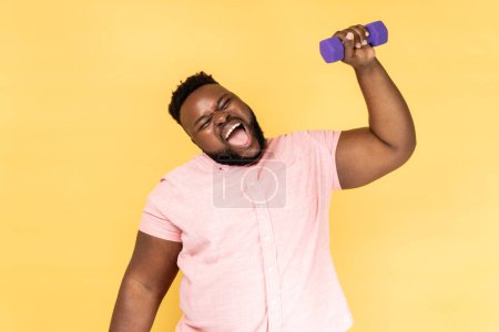 Photo for Portrait of funny positive bearded man wearing pink shirt standing and raised arm with dumbbell, making effort raising heavy sport equipment. Indoor studio shot isolated on yellow background. - Royalty Free Image
