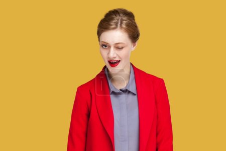 Foto de Portrait of positive flirting woman with red lips blinking her eyes with pleasure having happy expression, wearing red jacket. Indoor studio shot isolated on yellow background. - Imagen libre de derechos