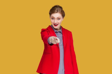 Foto de Portrait of happy excited woman with red lips standing pointing fore finger to camera, choosing you, expressing positive emotions, wearing red jacket. Indoor studio shot isolated on yellow background. - Imagen libre de derechos