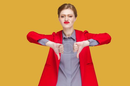 Photo for Portrait of sad upset young adult woman with red lips standing and showing thumbs down, dislike service, demonstrate disapprove, wearing red jacket. Indoor studio shot isolated on yellow background. - Royalty Free Image