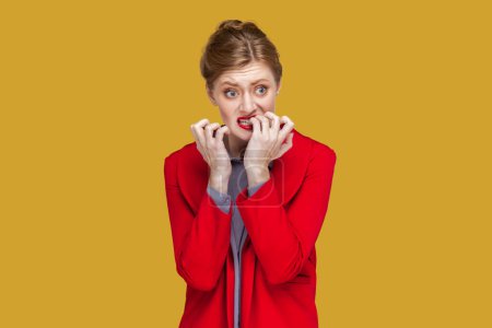 Photo for Portrait of nervous stressed woman with red lips standing biting her fingernails, looking away, being worried and depressed, wearing red jacket. Indoor studio shot isolated on yellow background. - Royalty Free Image