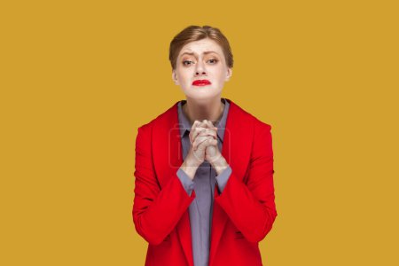 Téléchargez les photos : Portrait of hopeful woman with red lips standing with hands together, pleading, asking to forgive her, looking at camera, wearing red jacket. Indoor studio shot isolated on yellow background. - en image libre de droit
