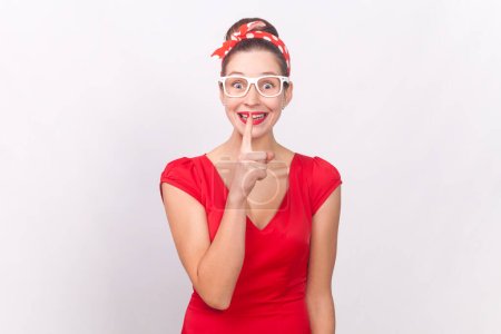 Photo for Portrait of funny happy smiling woman wearing red dress and head band keeps hand near lips, asking to keep secret, private information. Indoor studio shot isolated on gray background. - Royalty Free Image