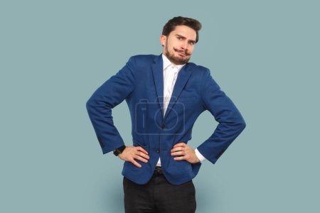 Photo for Portrait of funny angry man with mustache standing with hands on hips, looking at camera with frowning face, wearing official style suit. Indoor studio shot isolated on light blue background. - Royalty Free Image