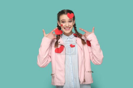 Téléchargez les photos : Portrait of romantic smiling teenager girl with braids wearing pink jacket standing pointing at little red heart on her body, expressing happiness. Indoor studio shot isolated on green background. - en image libre de droit