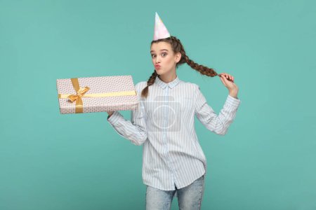 Photo for Flirting delighted attractive teenager girl with braids wearing striped shirt and party cone, holding her present on birthday, sending air kisses. Indoor studio shot isolated on green background. - Royalty Free Image