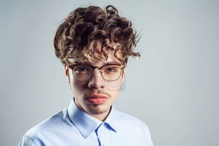 Photo for Portrait of serious young adult man wearing blue shirt and eyeglasses, with curly hairstyle, looking at camera with strict expression. Indoor studio shot isolated on gray background. - Royalty Free Image