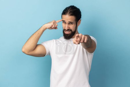 Téléchargez les photos : You are nuts. Portrait of man with beard showing stupid gesture and pointing to camera, accusing dumb insane suggestion, crazy plan for idiots. Indoor studio shot isolated on blue background. - en image libre de droit