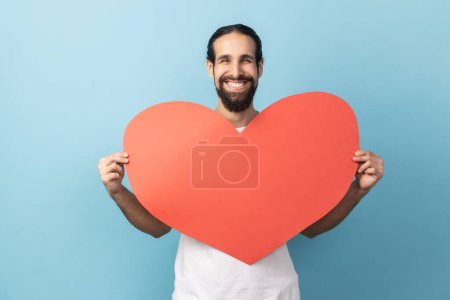 Photo for Portrait of delighted man with beard wearing white T-shirt holding big red heart in hands, looking at camera with positive, expressing love. Indoor studio shot isolated on blue background. - Royalty Free Image