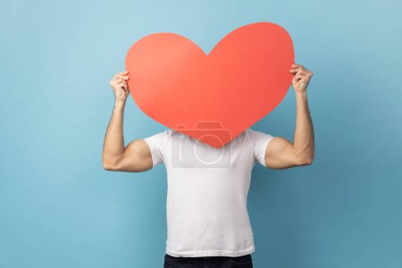 Photo for Portrait of man wearing white T-shirt hiding behind big red paper heart, holding symbol of love care, cardiac issues, demonstrating affection. Indoor studio shot isolated on blue background - Royalty Free Image