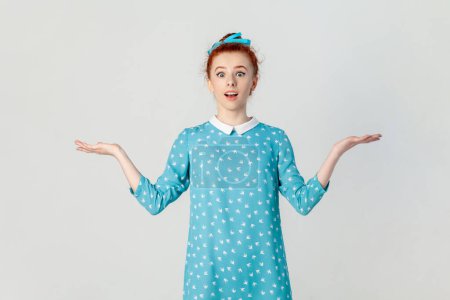 Photo for Portrait of shocked confused ginger woman standing with spread hands, looking at camera with big eyes, dosen't know how to react, wearing blue dress. Indoor studio shot isolated on gray background. - Royalty Free Image