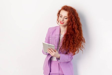 Téléchargez les photos : Portrait d'une joyeuse femme aux cheveux roux aux cheveux ondulés, debout et utilisant une tablette, faisant son travail en ligne, tapant, portant un costume lilas. Studio intérieur tourné isolé sur fond gris. - en image libre de droit