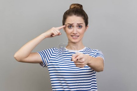 Foto de You are idiot. Woman wearing striped T-shirt showing stupid gesture and pointing to camera, blaming for insane plan, crazy idea, dumb suggestion. Indoor studio shot isolated on gray background. - Imagen libre de derechos