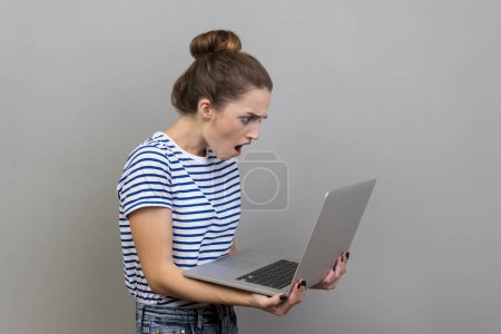 Photo for Side view of shocked woman wearing striped T-shirt working on laptop with surprised expression, freelance work, looking at display. Indoor studio shot isolated on gray background. - Royalty Free Image