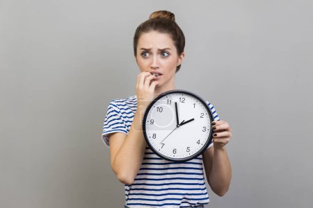 Photo for Portrait of woman wearing striped T-shirt holding wall clock in hands, biting nails, looking at camera with nervous expression, deadline. Indoor studio shot isolated on gray background. - Royalty Free Image