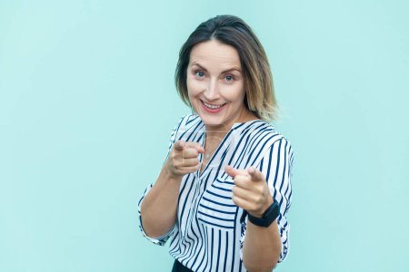 Photo for Portrait of satisfied attractive joyful middle aged woman wearing striped shirt pointing to camera with fingers, choosing you, making choice. Indoor studio shot isolated on light blue background. - Royalty Free Image