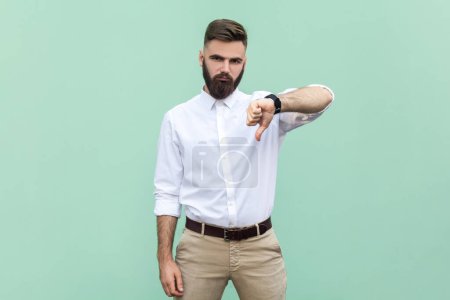 Photo for Bearded businessman wearing shirt criticizing bad quality with thumbs down displeased grimace, showing dislike gesture, expressing disapproval. Indoor studio shot isolated on light green background. - Royalty Free Image