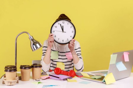 Photo for Woman office worker covered with sticky notes sitting at workplace hiding face behind big wall clock, schedule and business meeting appointment. Indoor studio studio shot isolated on yellow background - Royalty Free Image