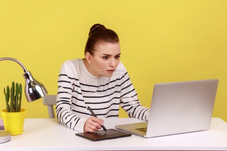 Téléchargez les photos : Femme sérieuse occupée concentrée travaillant sur une tablette graphique, dessinant des croquis, assise sur le lieu de travail avec un ordinateur portable regardant l'affichage. Studio intérieur tourné isolé sur fond jaune. - en image libre de droit