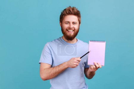 Photo for Portrait of bearded man pointing empty sheet and smiling at camera, showing paper notebook template, mock up blank space for business idea, message. Indoor studio shot isolated on blue background. - Royalty Free Image