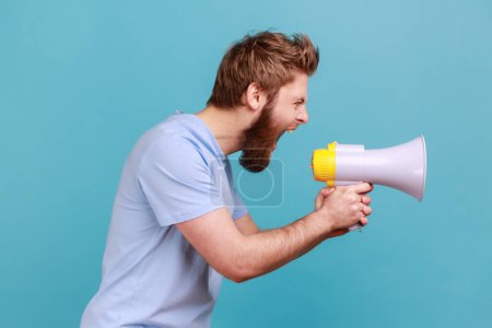 Photo for Side view of bearded man holding megaphone near mouth, loudly speaking, screaming, making announcement, paying attention at social problems. Indoor studio shot isolated on blue background. - Royalty Free Image