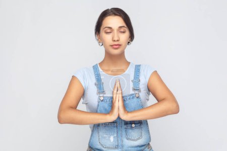Photo for Portrait of calm relaxed young adult woman wearing denim overalls standing palm hands and doing yoga meditation with closed eyes. Indoor studio shot isolated on gray background. - Royalty Free Image