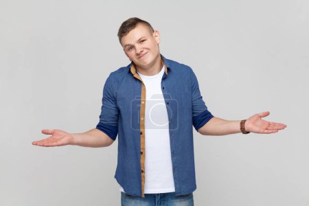 Photo for Portrait of open hearted generous teenager boy wearing blue shirt sharing opening hands looking at camera with kind smile, greeting and regaling. Indoor studio shot isolated on gray background. - Royalty Free Image