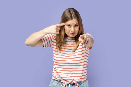 Foto de Retrato de una mujer rubia disgustada con una camiseta a rayas sosteniendo el dedo cerca de la sien y señalándote, mostrando un gesto estúpido, burlándose. Estudio interior plano aislado sobre fondo púrpura. - Imagen libre de derechos