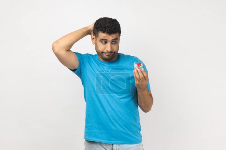 Photo for Portrait of confused puzzled handsome unshaven man wearing blue T- shirt standing with little gift box, having pensive and thoughtful expression. Indoor studio shot isolated on gray background. - Royalty Free Image