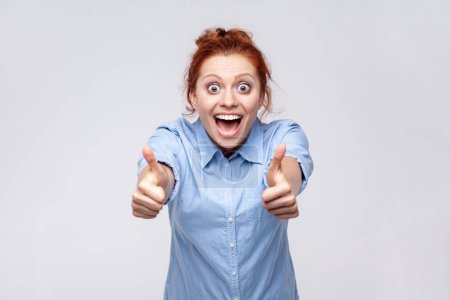Photo for Portrait of positive redhead woman wearing blue shirt showing thumbs up, looking at camera with open mouth, surprised with excellent result. Indoor studio shot isolated on gray background. - Royalty Free Image